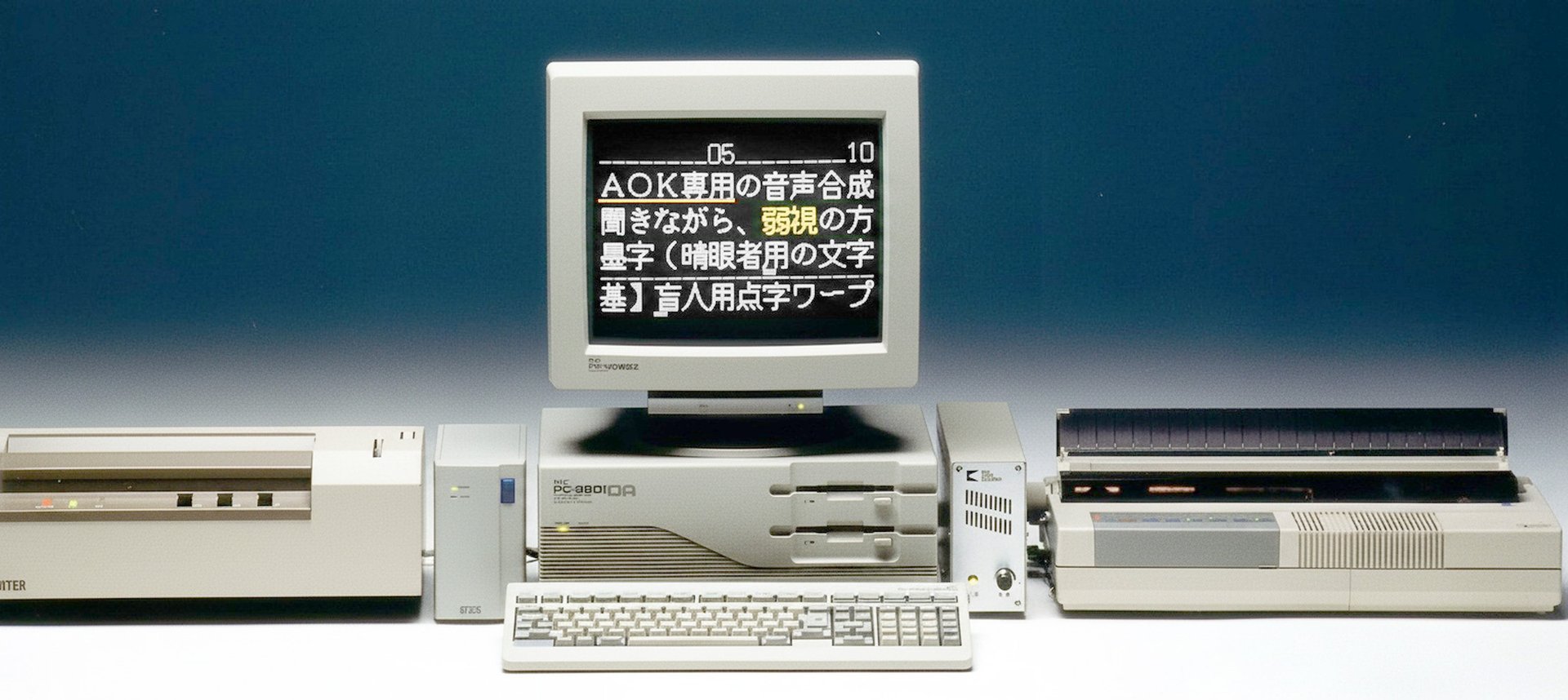 会社設立当時のAOK点字日本語ワープロのシステム写真。中央のパソコン(PC-9801)の画面にAOK専用の音声合成の説明文が表示され、キーボードと音声合成装置、左右に点字プリンターなどの周辺機器が並んでいる
